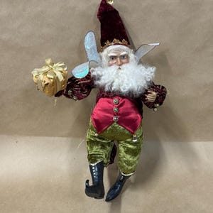 May include: A whimsical fairy figurine with a long, burgundy velvet hat and matching jacket. The fairy has a white beard, green velvet pants, and black boots. It holds a gold gift box, perfect for holiday decor.