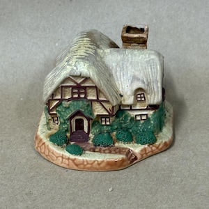 May include: A miniature ceramic cottage with a thatched roof, brown trim, and green ivy. The cottage has a chimney and small windows. The base is a light brown with a stone path leading to the front door. A charming decorative piece.