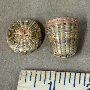 May include: A miniature woven basket with a separate circular lid, crafted from light tan natural fibers. The basket features a slightly flared rim with a pinkish-tan accent band. A wooden ruler shows the scale in inches at the bottom.