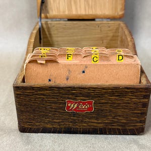 May include: A dark brown wooden index card box with a hinged lid and a red and gold Weis label on the front. Inside, the box contains several tan paper index cards with yellow alphabetical tabs labeled with letters including F, C, and D.