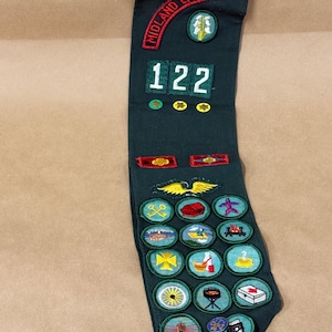 May include: Dark green sash with embroidered patches. The top reads "MIDLAND EMPIRE" in red. Features the number "122" and various colorful patches with images like a wheel, a campfire, and a first aid kit.