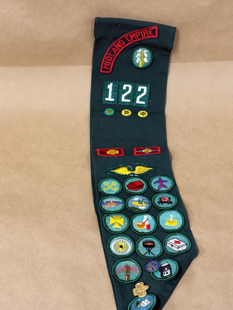 May include: Dark green sash with embroidered patches. The top reads "MIDLAND EMPIRE" in red. Features the number "122" and various colorful patches with images like a wheel, a campfire, and a first aid kit.