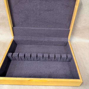May include: Open wooden silverware storage box with a light brown exterior and a dark gray felt interior. The box has a sectioned area for holding silverware. The box is open, revealing the interior.