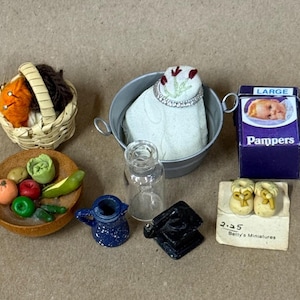 May include: A collection of miniature items including a woven basket with yarn, a metal bucket containing a white fabric piece, a Pampers box, a wooden bowl with fruit, a blue pitcher, a glass bottle, a black object, and baby shoes.
