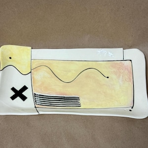 May include: A rectangular ceramic tray with a white base, featuring a yellow-tinted section on the left, a central yellow and peach-toned rectangular area with a black wavy line, a bold black X, and a series of horizontal black stripes.