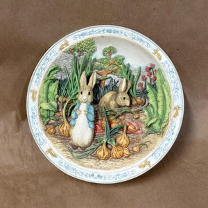 May include: A round decorative plate featuring a three-dimensional relief of two rabbits in a garden. One rabbit wears a blue jacket, standing among green leafy plants and brown onions. The plate rim has a blue floral pattern and small gold rabbits.