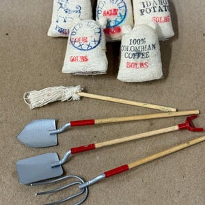 May include: A collection of five miniature burlap-style sacks labeled with text like Idaho Potato 50LBS and 100% Colombian Coffee 50LBS, alongside a miniature mop and four garden tools including a shovel, spade, and pitchfork with wooden handles.