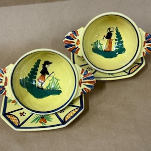 May include: Two yellow ceramic bowls with matching saucers. Each bowl features a hand-painted scene of a person and a tree. The saucers have a geometric design with orange, blue, and green accents. The bowls have handles with a fan-like design.