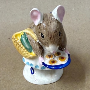 May include: A small ceramic mouse figurine with brown fur and pink-lined ears. The mouse wears a white apron with blue dots, carries a yellow woven basket containing a green vegetable, and holds a white plate with four small round cookies.