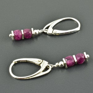 Könnte beinhalten: Ein Paar silberne Ohrringe mit Klappbügeln. Jeder Ohrring hat drei facettierte, tiefrosa Edelsteine, getrennt durch silberne Akzente. Die Ohrringe haben ein schlichtes, elegantes Design.