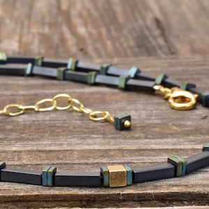 Könnte beinhalten: Eine Halskette mit dunkelgrauen, rechteckigen Perlen mit goldenen und türkisfarbenen Akzenten. Die Kette hat eine goldfarbene Kette und einen Verschluss. Das Design ist modern und geometrisch.