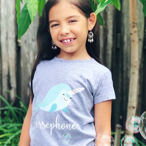 K&ouml;nnte beinhalten: Ein graues T-Shirt mit einem hellblauen Narwal-Motiv und dem Namen "Persephone" auf der Vorderseite. Das Shirt wird von einer jungen Person mit langen dunklen Haaren und silbernen Ohrringen getragen.