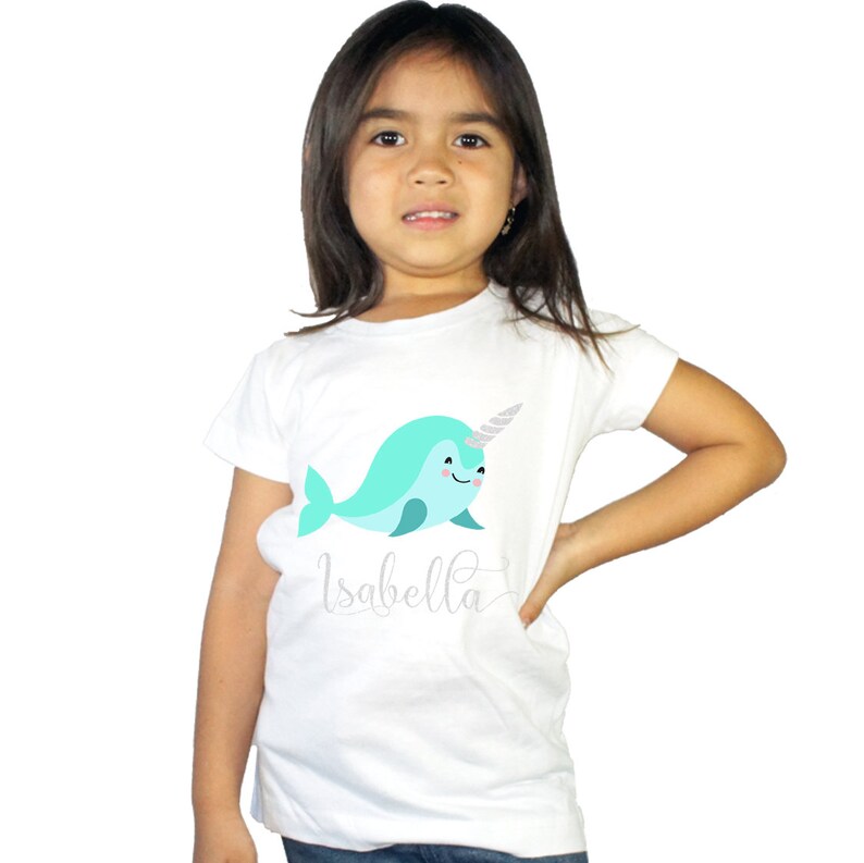 K&ouml;nnte beinhalten: Wei&szlig;es T-Shirt mit einem hellblauen Narwal-Motiv und dem Namen "Isabella" in silbernen Glitzerbuchstaben.