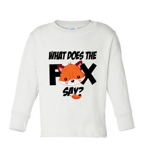 Może przedstawiać: Biała koszulka z długim rękawem z kresk&oacute;wkowym lisem w kolorze pomarańczowym i brązowym. Tekst "WHAT DOES THE FOX SAY?" jest wydrukowany nad i pod ilustracją lisa w kolorze czarnym.