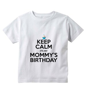 May include: White t-shirt with black text that reads "Keep Calm It's My Mommy's Birthday". A small blue cupcake with a lit candle is above the text.