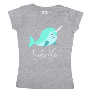 K&ouml;nnte beinhalten: Graues T-Shirt mit einem hellblauen Narwal mit einem silbernen Horn und dem Namen "Isabella" in wei&szlig;er Kursivschrift.