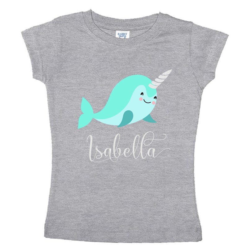 K&ouml;nnte beinhalten: Graues T-Shirt mit einem hellblauen Narwal mit einem silbernen Horn und dem Namen "Isabella" in wei&szlig;er Kursivschrift.