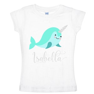 K&ouml;nnte beinhalten: Wei&szlig;es T-Shirt mit einem t&uuml;rkisfarbenen Narwal mit einem silbernen Glitzerhorn und dem Namen "Isabella" in silbernen Glitzerbuchstaben.