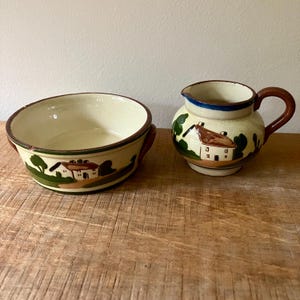May include: A set of two vintage ceramic pieces with a cottage scene painted on them. The set includes a small pitcher with a brown handle and a round bowl with a brown rim.
