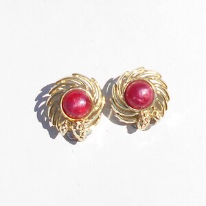 May include: A pair of gold earrings with a red gemstone in the center of each earring. The earrings have a swirl design.
