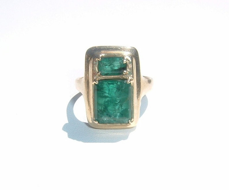 May include: A gold ring featuring two emerald green rectangular gemstones set in a gold frame.