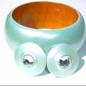 May include: A set of two light blue wooden earrings with clear rhinestone accents. The earrings are round and have a slightly raised center.
