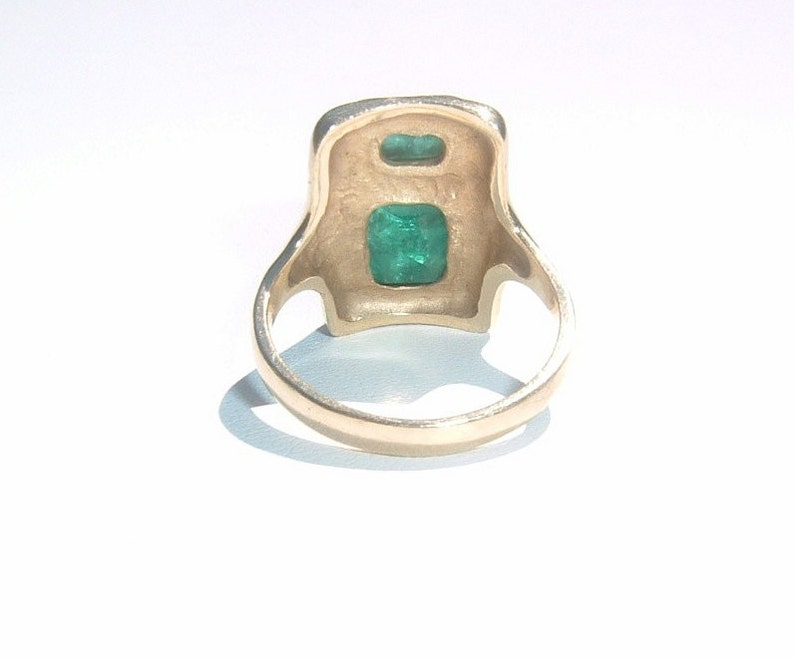 May include: A gold ring with two emerald green gemstones set in a square-shaped design.