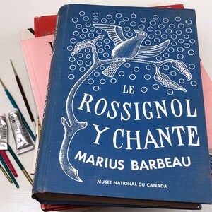 May include: A blue book with white text and illustrations. The title is "Le Rossignol Y Chante" by Marius Barbeau. The book is part of a stack of books on a white surface.
