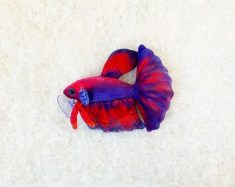 May include: A plush toy shaped like a red and purple betta fish. The fish has a large, flowing tail fin and a small, round body.