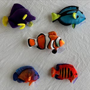 May include: Five colorful plush fish toys with suction cups. The fish are various colors, including purple, blue, orange, and red, with yellow and black accents. The toys are designed to stick to smooth surfaces.