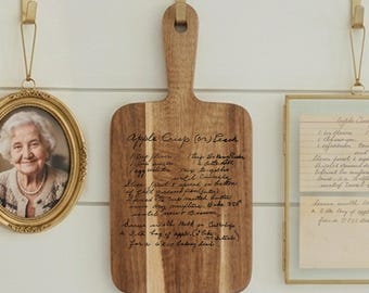 May include: A rectangular wooden cutting board with a handle, featuring a dark brown wood grain and black engraved cursive text that reads Apple Crisp (or) Peach, followed by a handwritten recipe including ingredients and baking instructions.