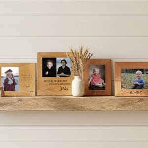 May include: Four rectangular light-colored wooden picture frames displayed on a shelf. The frames contain various photographs of individuals and children, with engraved text including "FROM LITTLE STEPS TO CAP AND GOWN", "MYLEE", and "BLAKE".