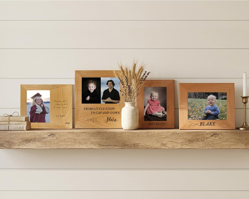 May include: Four rectangular light-colored wooden picture frames displayed on a shelf. The frames contain various photographs of individuals and children, with engraved text including "FROM LITTLE STEPS TO CAP AND GOWN", "MYLEE", and "BLAKE".