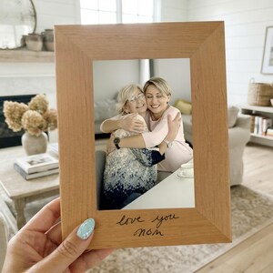 May include: A rectangular light brown wooden picture frame with the handwritten text "love you mom" engraved on the bottom border. The frame holds a photograph of two people embracing, with one person wearing glasses and a patterned top and the other in pink.