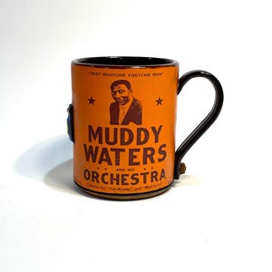 May include: Orange mug featuring a portrait of Muddy Waters and the text "Muddy Waters and his Orchestra." The mug has a black handle and rim, with the quote "That Hootchie Cootchie Man" at the top. The mug is decorated with stars.