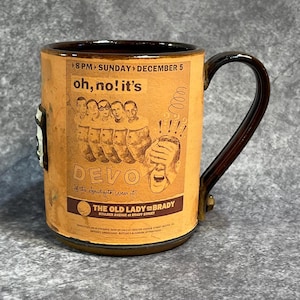 May include: A ceramic mug with a brown handle and rim, featuring a vintage-style poster design. The poster reads "oh, no! it's DEVO" with a graphic of band members and the text "THE OLD LADY on BRADY".