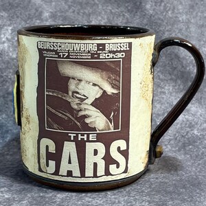 May include: A ceramic mug with a dark brown handle and rim. The mug features a vintage poster design for "The Cars" band, with text including "BEURSSCHOUWBURG - BRUSSEL" and "17 NOVEMBRE". The mug has a cream-colored background.