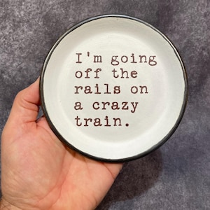 May include: A small, round, white ceramic dish with a black rim. The text "I'm going off the rails on a crazy train" is printed in brown, centered on the dish. The dish is held in a hand against a gray background.