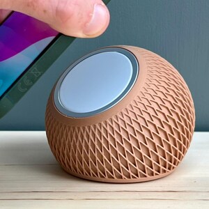 May include: A spherical, peach-colored charging dock with a textured, cross-hatched lattice pattern on its exterior. The device features a circular, light-colored magnetic charging surface on its angled top, designed to hold a smartphone in place.