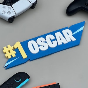 May include: A blue sign with the name "OSCAR" in white block letters, and a yellow "#1" symbol. The sign is surrounded by white, black, and blue game controllers, suggesting a gaming theme.