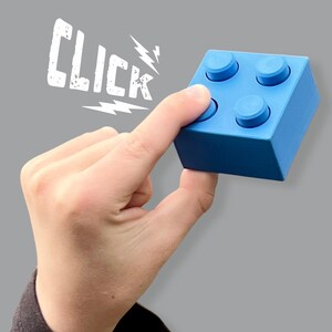 May include: A blue building block with four studs is held by a hand. The word "CLICK" is written in white, with lightning bolt graphics. The block is a solid, square shape.