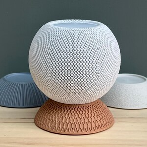 May include: A spherical white smart speaker with a textured lattice exterior sits atop a terracotta-coloured circular base with a matching lattice pattern. Two additional circular bases, one blue with vertical ridges and one white with a rough texture, are placed behind.