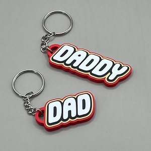 May include: Two keychains with the words "DADDY" and "DAD" in white block letters with a red border. The keychains have a silver key ring attached.