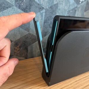 May include: A black docking station with a light blue interior, designed for a gaming console. A finger is shown lifting a section of the docking station. The docking station is on a wooden surface.