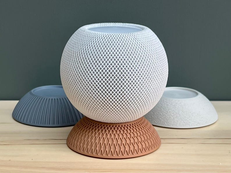 May include: A spherical white smart speaker with a textured lattice exterior sits atop a terracotta-coloured circular base with a matching lattice pattern. Two additional circular bases, one blue with vertical ridges and one white with a rough texture, are placed behind.