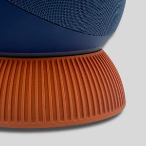 Puede incluir: Un altavoz inteligente azul marino y naranja. El altavoz tiene una parte superior de malla azul oscuro y se asienta sobre una base naranja y acanalada. La base tiene una forma redondeada con l&iacute;neas verticales. El altavoz es de dise&ntilde;o moderno.