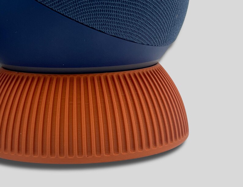 Puede incluir: Un altavoz inteligente azul marino y naranja. El altavoz tiene una parte superior de malla azul oscuro y se asienta sobre una base naranja y acanalada. La base tiene una forma redondeada con l&iacute;neas verticales. El altavoz es de dise&ntilde;o moderno.