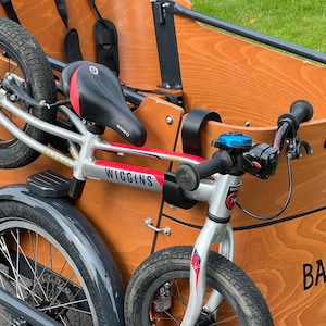 May include: A Babboe cargo bike with a wooden cargo box and a silver frame. The bike has black tires, a black seat with red accents, and a blue bell. The brand name "Wiggins" is visible on the frame.