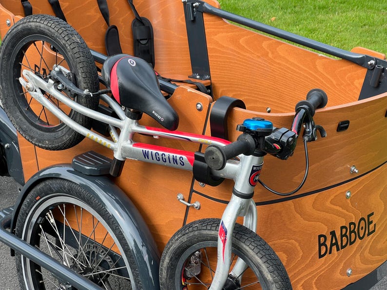 May include: A Babboe cargo bike with a wooden cargo box and a silver frame. The bike has black tires, a black seat with red accents, and a blue bell. The brand name "Wiggins" is visible on the frame.