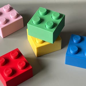 May include: A collection of colorful building block toys in pink, green, yellow, blue, and red. Each block has four raised circular studs on top. The blocks are arranged on a light gray surface.
